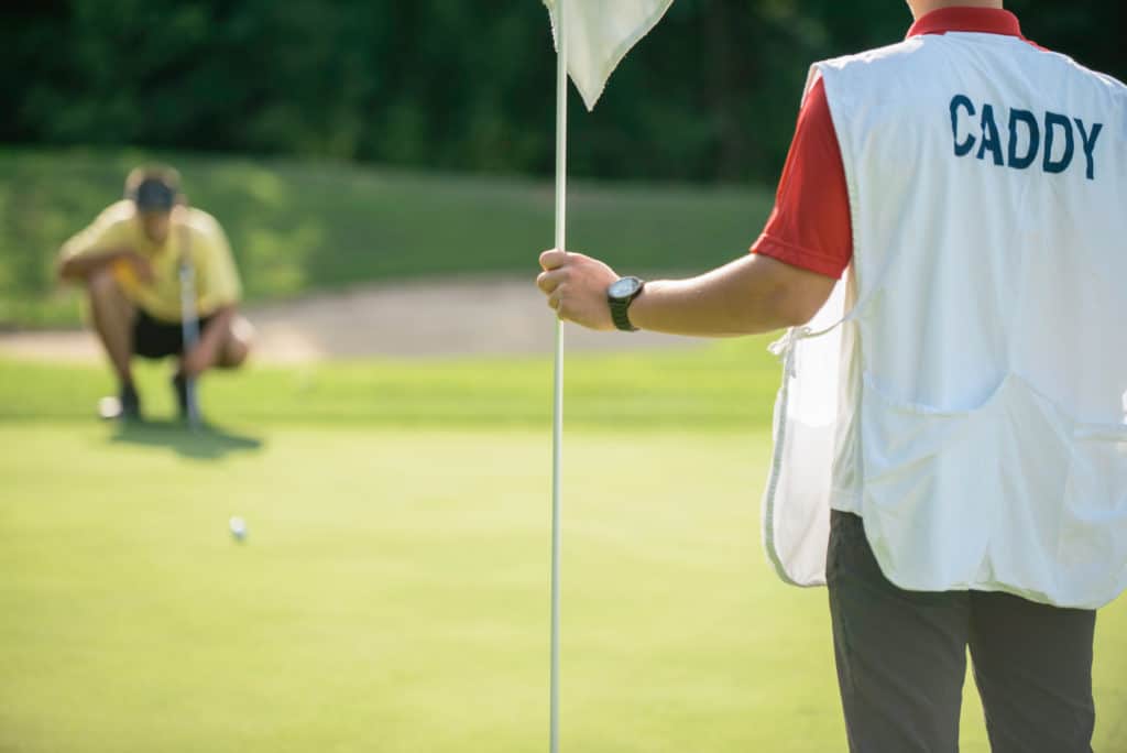 Golfer putting from long distance, reading green, caddy holding golf flag