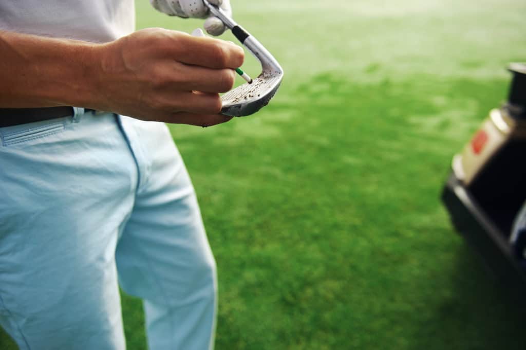 golfer removes dirt and sand from grooves of iron club with a tee peg