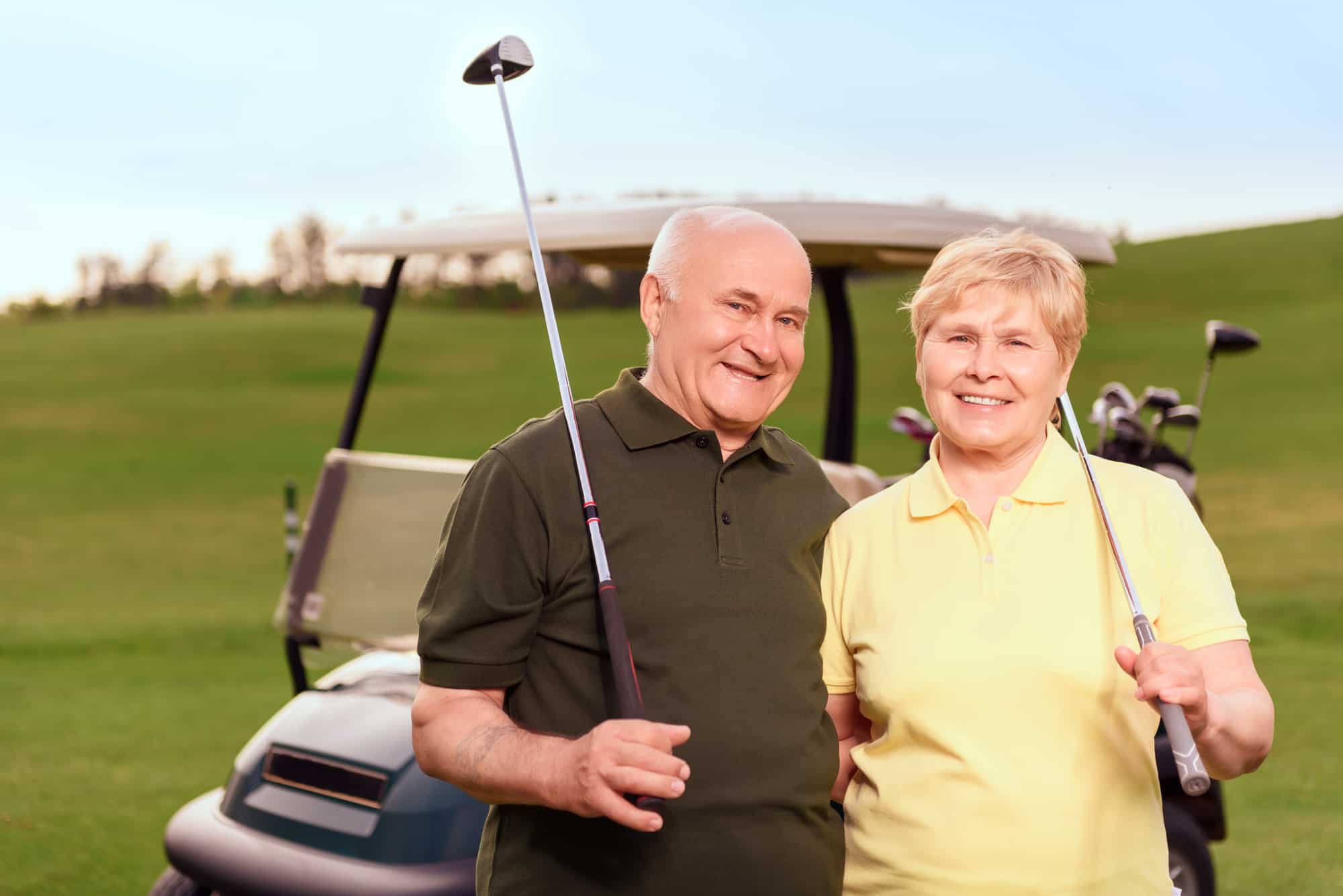 Satisfied with game. Two lovely senior people standing with golf clubs on background of cart on course.