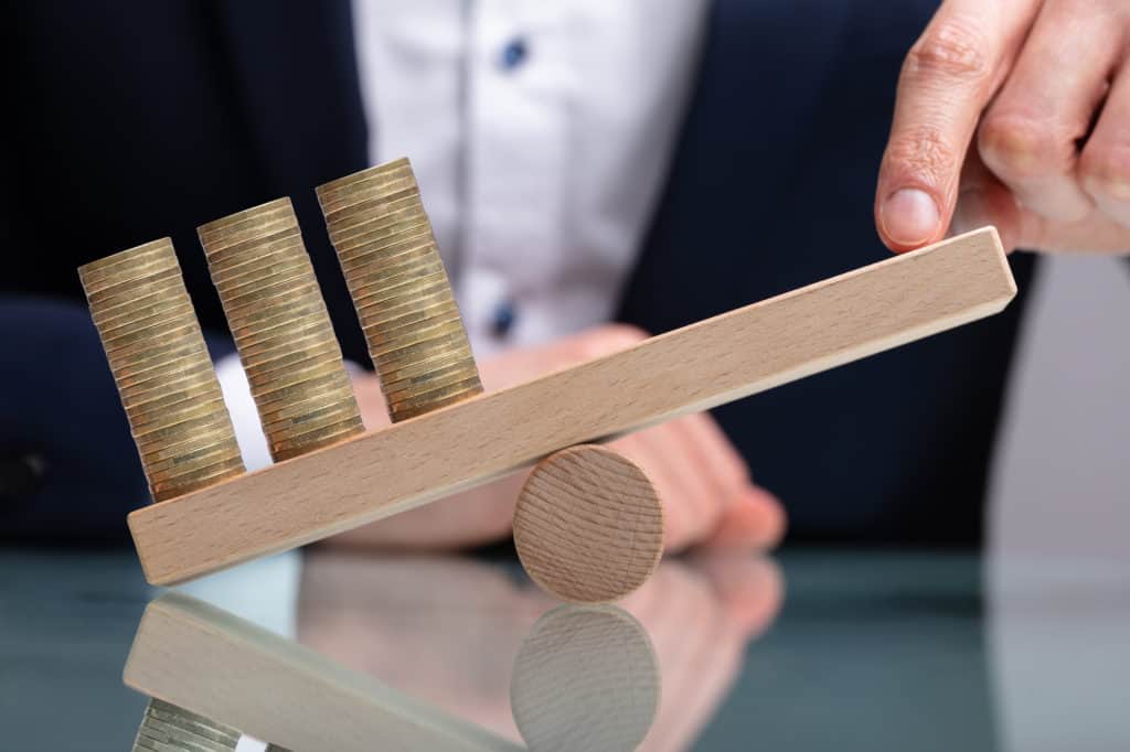 Businessman Balancing Stacked Coins With Finger On Wooden Seesaw Over Reflective Desk