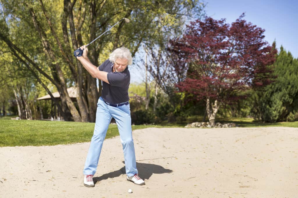 Old golf player swinging golf club in sand bunker.