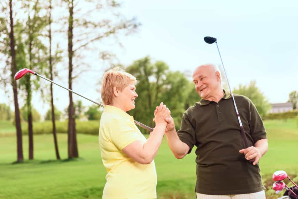 One team. Smiling senior man and woman standing on course holding golf clubs and their hands together