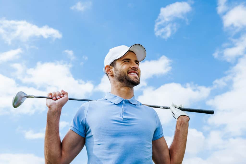 Looking towards success. Low angle view of young happy golfer holding driver and smiling with blue sky as background