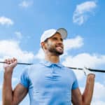 Looking towards success. Low angle view of young happy golfer holding driver and smiling with blue sky as background