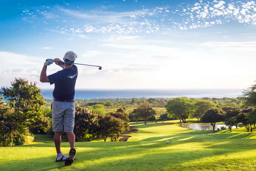 Man hitting golf ball down hill towards ocean and horizon