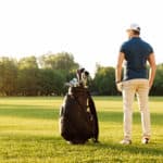 Back view of a young man standing on a green field with golf bag