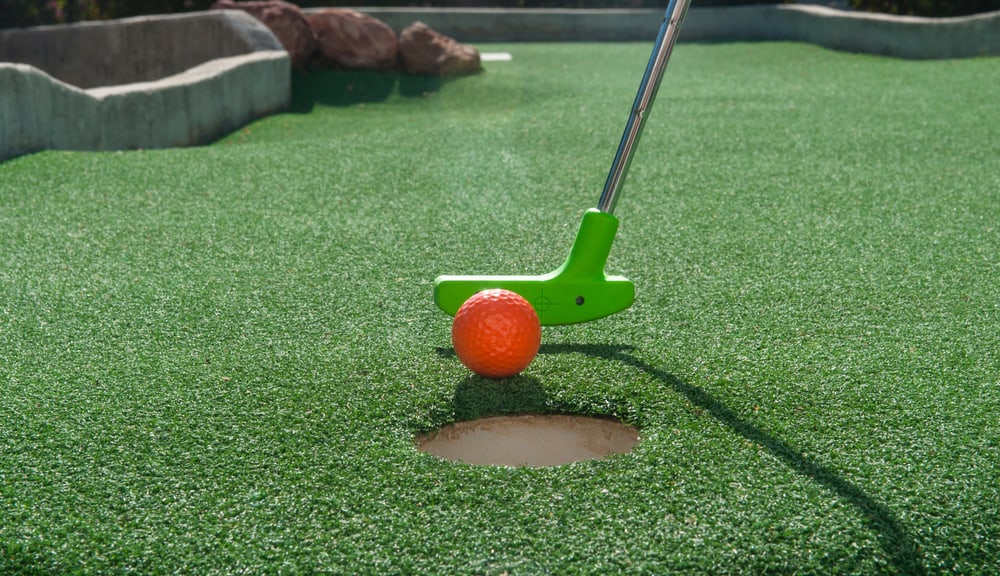 Green mini golf putter ready to tap an orange ball into the hole on green turf