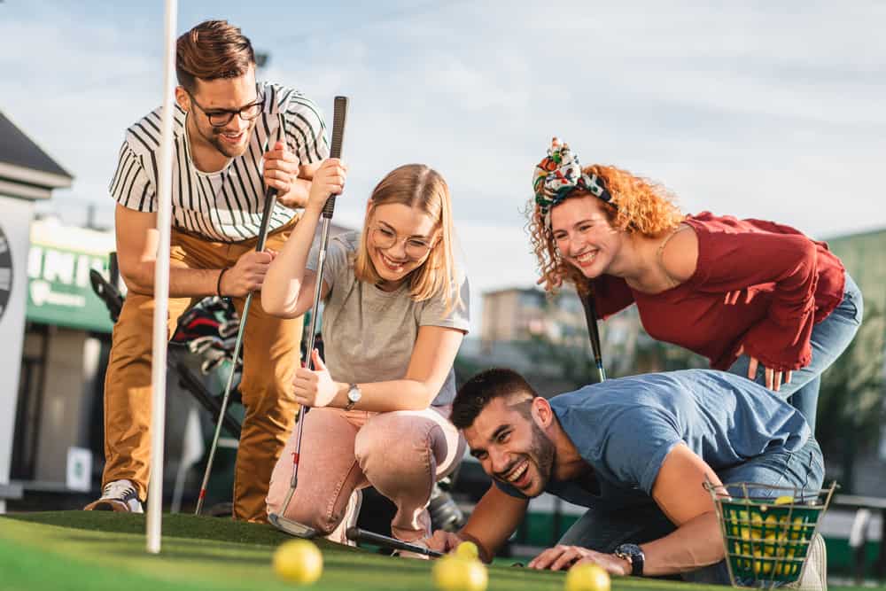 Group of smiling friends enjoying together playing mini golf in the city