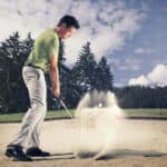 Male golf player in green shirt and grey pants hitting golf ball out of a sand trap