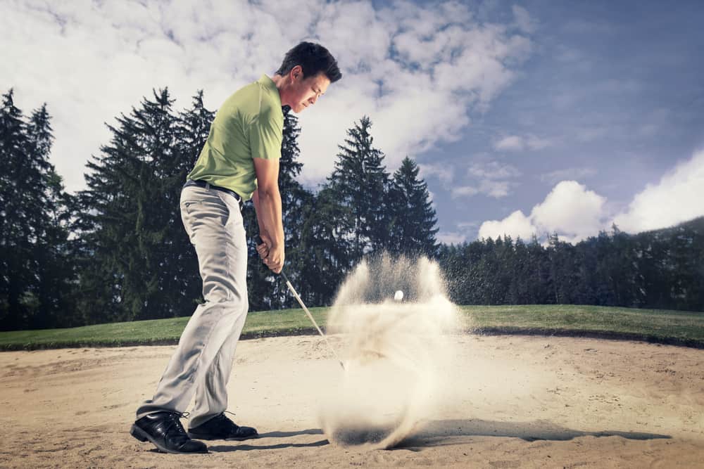 Male golf player in green shirt and grey pants hitting golf ball out of a sand trap