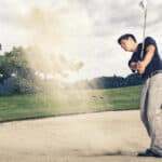Male golfer in blue shirt and grey pants hitting golf ball out of a sand trap