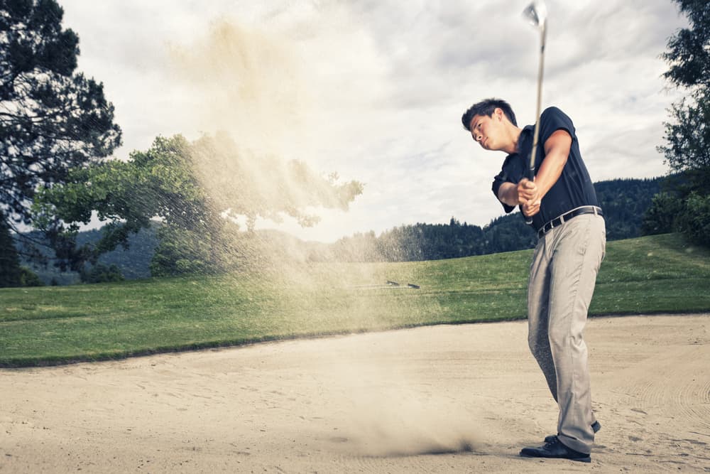 Male golfer in blue shirt and grey pants hitting golf ball out of a sand trap