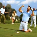 Happy golfer kneeling at hole with raised fists after putting in golf ball to the hole