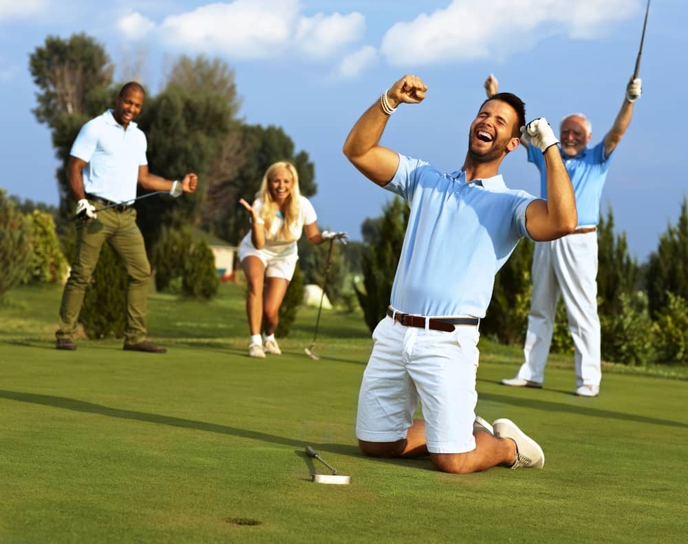 Happy golfer kneeling at hole with raised fists after putting in golf ball to the hole