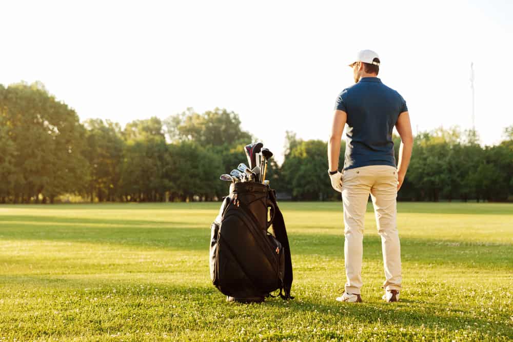 Back view of a young man standing on a green field with golf bag